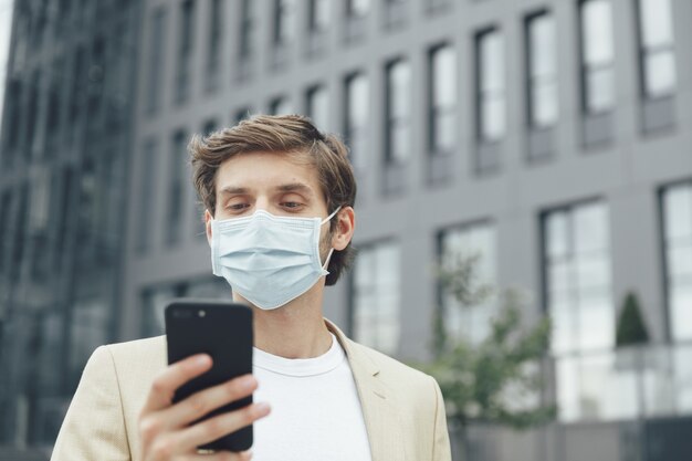 Attractive happy man in protective mask and business suit using modern smartphone outdoors