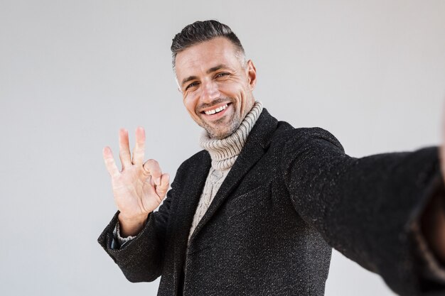 Attractive happy man wearing coat standing isolated over gray wall, taking a selfie, showing ok