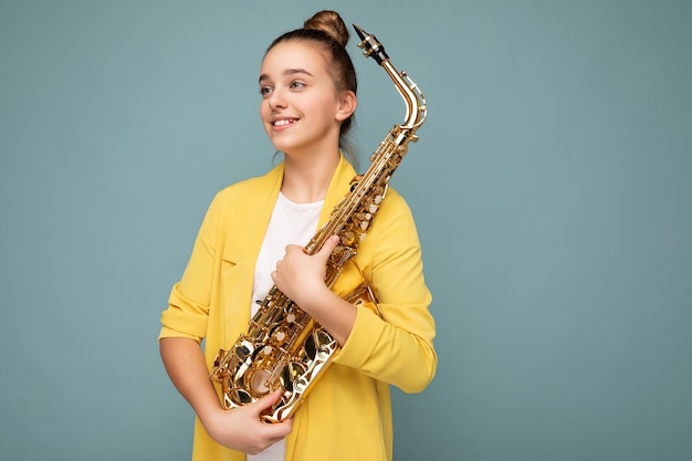 Attractive happy smiling brunette little female teenager wearing trendy yellow jacket standing isolated over blue background wall holding saxophone looking to the side. Empty space, copy space