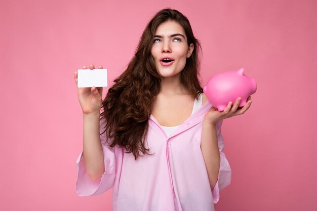 Attractive happy surprised young brunette woman wearing shirt isolated on pink background with empty