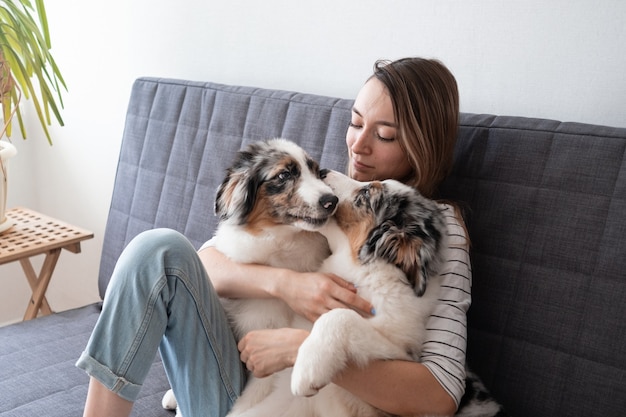 매력적인 행복한 여성이 귀여운 호주 셰퍼드 블루 멀 강아지 두 마리를 껴안고 키스합니다. 소파에 앉아. 애완 동물 관리와 친절한 개념. 인간과 동물 사이의 사랑.