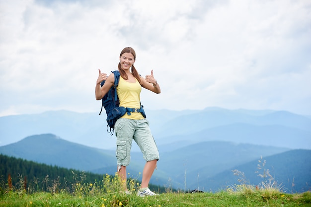 Attractive happy woman hiker hiking in mountain trail, walking on grassy hill, smiling and showing thumbs up, enjoying summer day