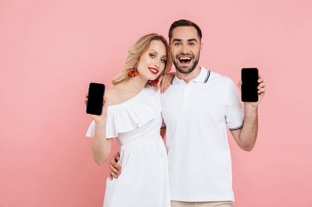 Attractive happy young couple standing together isolated over pink , showing blnk screen mobile phones