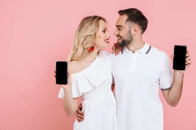 Attractive happy young couple standing together isolated over pink , showing blnk screen mobile phones