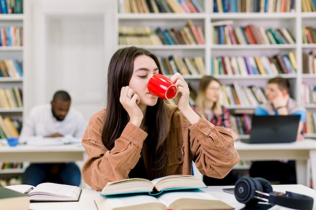 魅力的な幸せな若い女の子の学生、茶色のカジュアルシャツを着て、大学図書館で勉強して、たくさんの本を机に座って、コーヒーを飲む