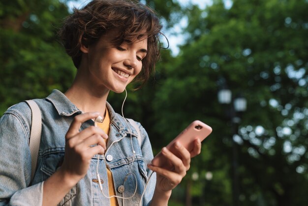 캐주얼 복장을 한 매력적인 어린 소녀는 공원에서 야외에서 시간을 보내고 이어폰으로 음악을 듣고 휴대폰을 들고 있습니다.