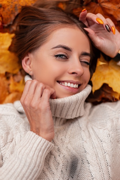 Attractive happy young woman winks looking at the camera. Portrait of a cheerful positive girl in a knitted white sweater in autumn orange foliage in the park. View from above.