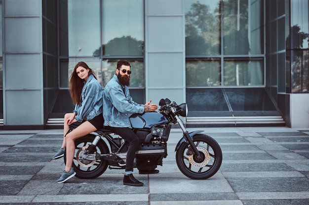 Attractive hipster couple - bearded brutal male in sunglasses and jeans jacket and his young sensual girl sitting back to back on a retro motorcycle against a skyscraper.