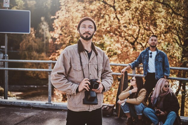 彼の友人がバックグラウンドで休んでいる間、眼鏡の魅力的なヒップスターは写真カメラで写真家のためにポーズをとっています。