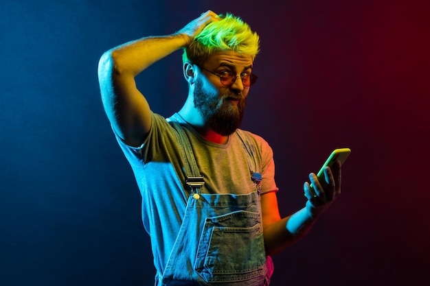 Attractive hipster guy holding smart phone in hands having puzzled and confused facial expression keeps hand on head looks at camera with frowning face Colorful neon light indoor studio shot