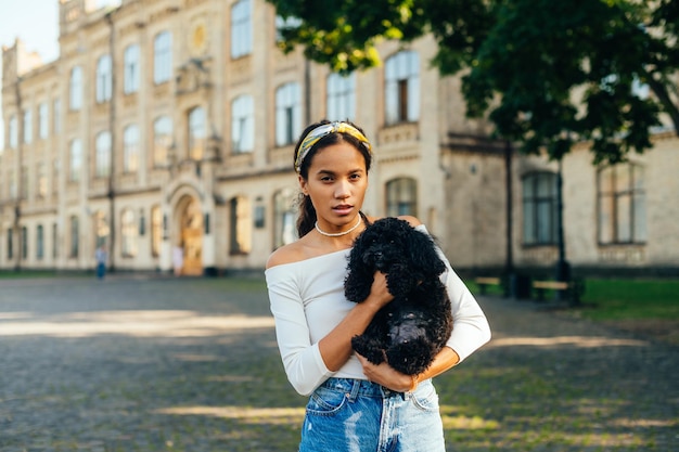 Attractive hispanic girl standing in the park on the background of old architecture with a small dog