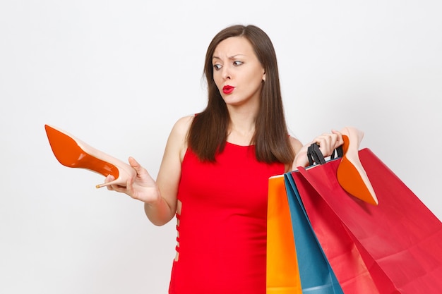 Attractive impressive glamour caucasian fashionable young brown-hair woman in red dress holding beige shoes with red sole, multi colored packets with purchases, shopping isolated on white background.