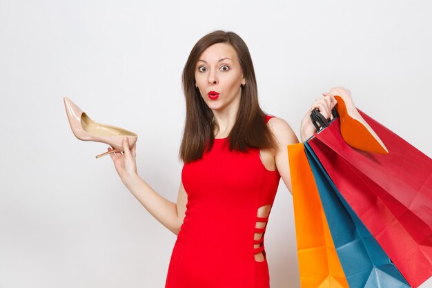 Attractive impressive glamour caucasian fashionable young brown-hair woman in red dress holding beige shoes with red sole, multi colored packets with purchases, shopping isolated on white background.