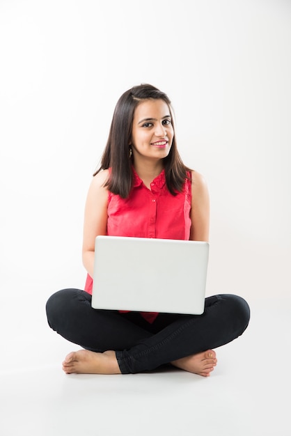 Attractive Indian Asian girl student studying on laptop computer with pile of books, sitting isolated over white floor