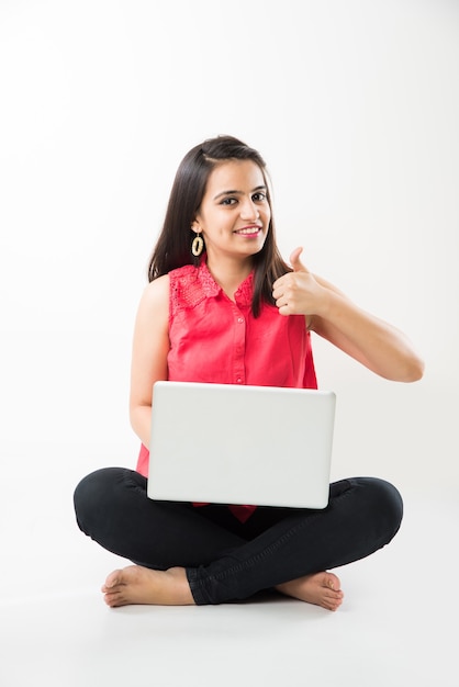 Attractive Indian Asian girl student studying on laptop computer with pile of books, sitting isolated over white floor