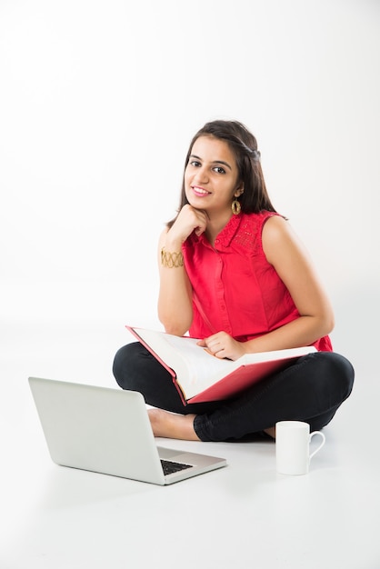 Attractive Indian Asian girl student studying on laptop computer with pile of books, sitting isolated over white floor