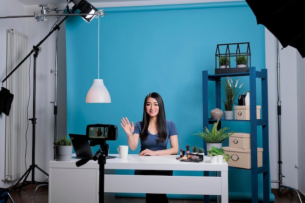 Attractive influencer greeting remote subscribes while recording make up tutorial advertising cosmetics product. Behind the scene of creative creator filming makeup tutorial using podcast equipment