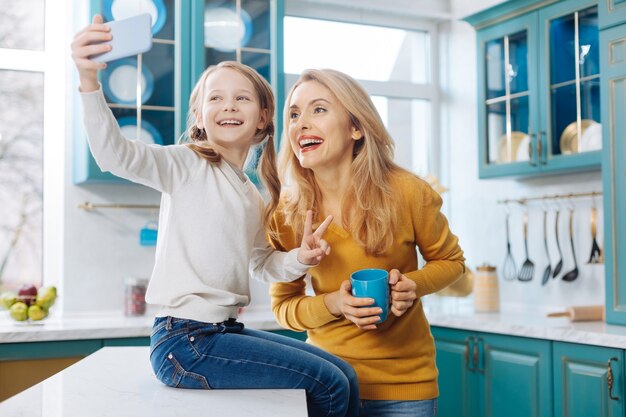 Привлекательная вдохновленная светловолосая стройная мать улыбается и держит чашку чая, пока ее дочь фотографирует