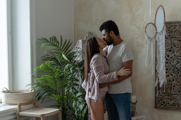 Attractive interracial couple in home clothes kiss and hug in\
boho home interior full length