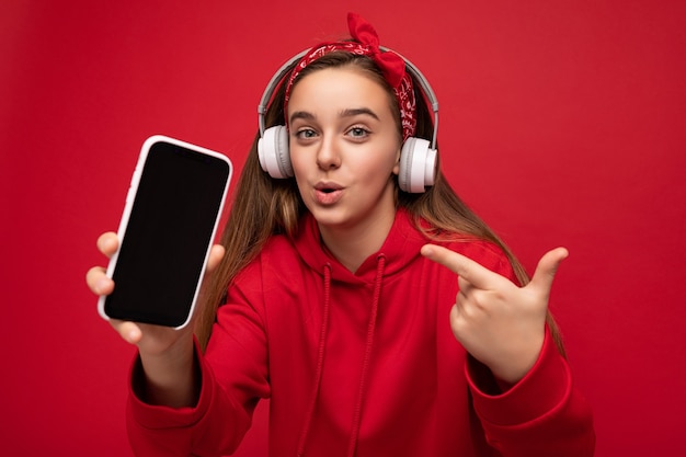 Attractive joyful amazed brunette girl wearing red hoodie isolated on red background