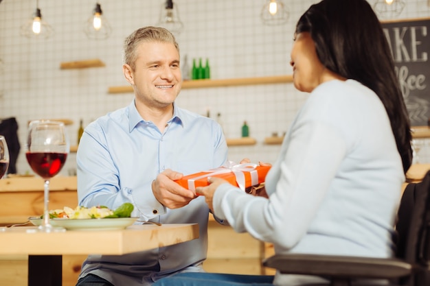 夕食を食べながら笑顔で彼の美しい陽気な障害者の女性にプレゼントを与える魅力的な楽しい金髪の男