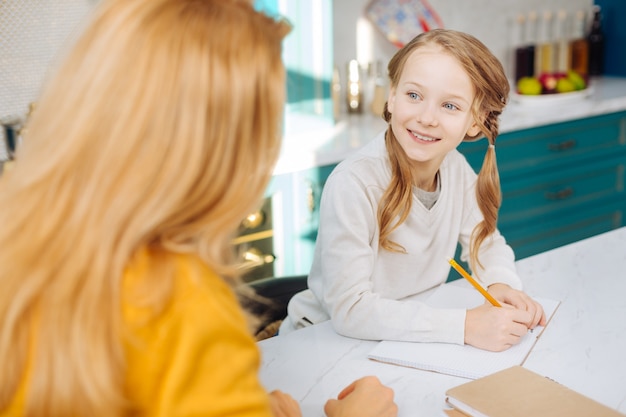 Attractive joyful fair-haired girl smiling and holding a pencil while looking at her mother and sitting at the table