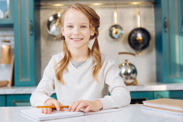 Attractive joyful fair-haired girl smiling and holding a pencil while sitting at the table with her notebooks