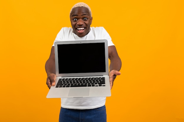 Attractive joyful surprised american man in white t-shirt holds out his hands with laptop with mockup on yellow background