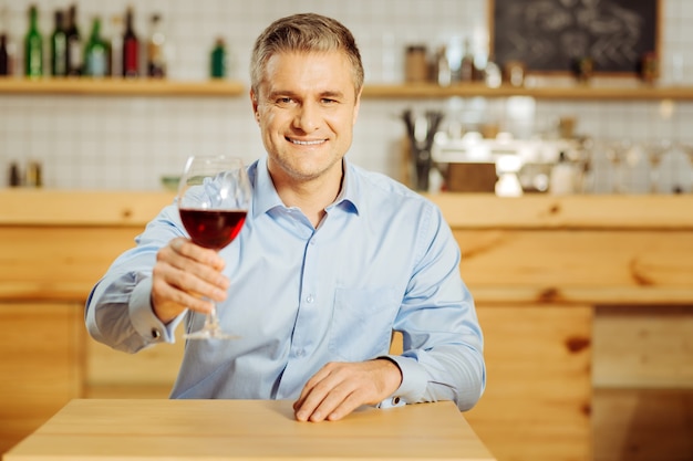 Привлекательный радостный хорошо сложенный мужчина улыбается и пьет вино во время отдыха в кафе