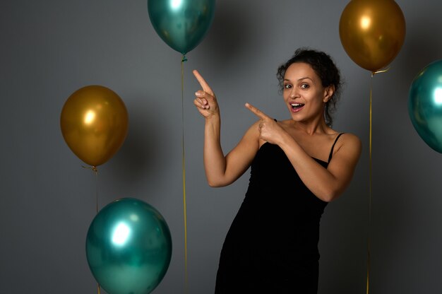 Attractive joyful woman in evening attire looks at camera and points on a gray background copy space decorated with golden green air balloons for Christmas, anniversary , New Year advertisement