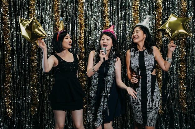 Attractive ladies cheerfully enjoying music at nightclub in
karaoke party. young girls dancing having fun playing with gold
star balloons. beautiful women wearing funny hats performing on
stage.