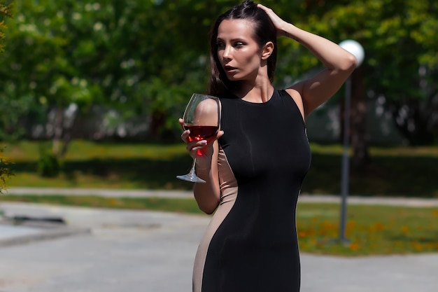 화창한 여름날 목을 만지고 레드 와인을 즐기는 검은색 우아한 드레스를 입은 매력적인 여성
