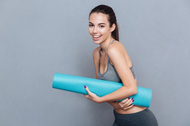 Attractive laughing sportswoman holding yoga mat isolated over grey background