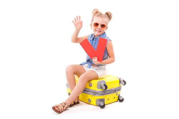 Attractive little girl in blue shirt, white shorts and sunglasses sit on the yellow suitcase and hold red cards