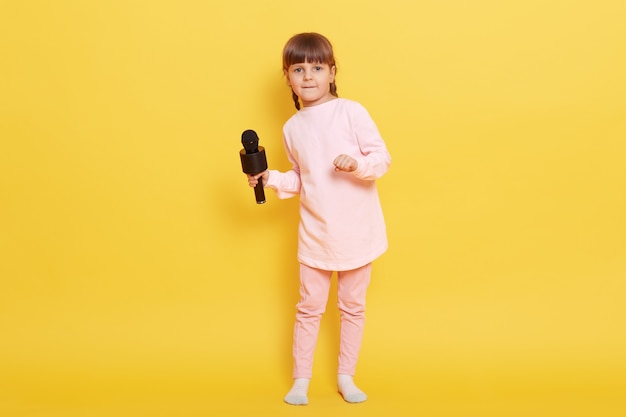 Attractive little girl with microphone wearing pink pale attire standing isolated over yellow background, charming vocalist sings song in karaoke, kid arranging concert.