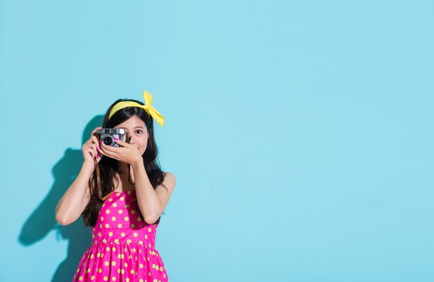 привлекательная милая девушка с помощью камеры в винтажном стиле фотографирует стоя на синем фоне и в летней милой одежде.