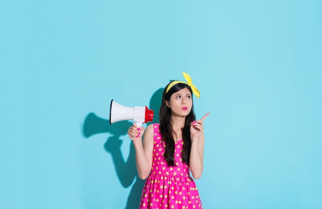 attractive lovely woman making pointing gesture looking at empty area and holding loudspeaker standing in blue background.