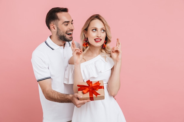 Attractive lovely young couple in love standing together isolated over pink , giving presents, holding fingers crossed for good luck, celebrating event
