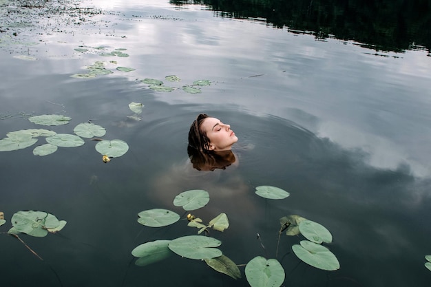 Attractive magic woman swimming in a lake among water lilies after rain. Art water portrait.