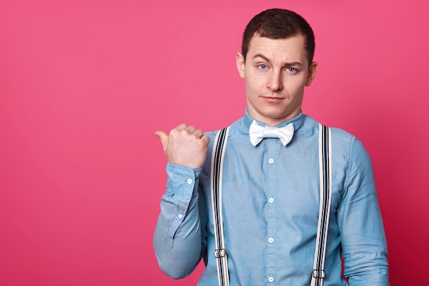 attractive male in blue shirt, suspenders and white bow tie, shows with thumb finger aside