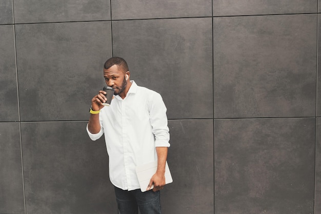 Attractive male in white shirt drinking a cup of coffee or tea\
taking a break from office work outdoor handsome stylish african\
american businessman or student looking at camera hold tablet in\
arms