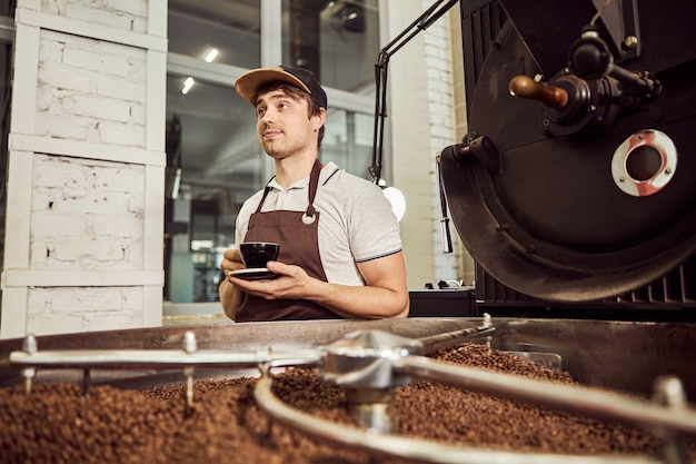 一杯のコーヒーを保持し、コーヒー豆を焙煎するための専門的な機器の近くに立っている間目をそらしているエプロンの魅力的な男性労働者