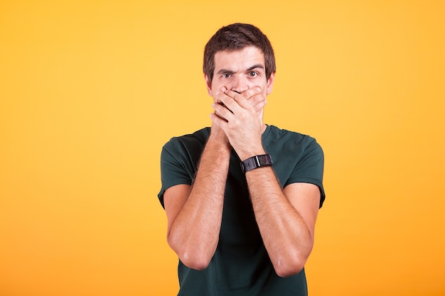 Attractive man in casual t-shirt covering his mouth in no talk sign on yellow background in studio photo.