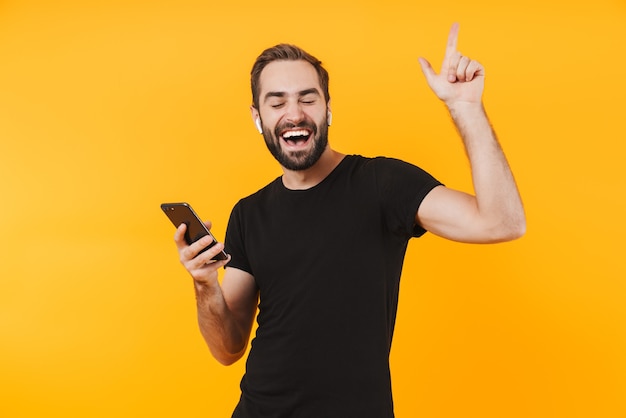 attractive man in earpods dancing and listening to music with cellphone isolated over yellow wall