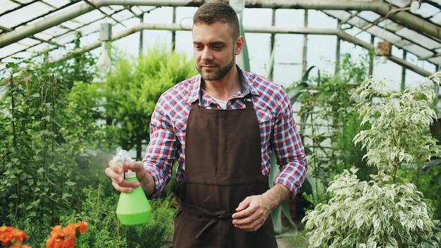 温室の庭の噴霧器と植物や花に水をまくエプロンの魅力的な男の庭師