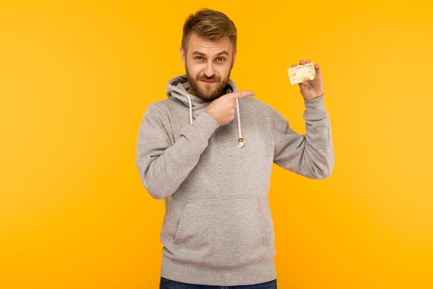 Привлекательный мужчина в серой толстовке с капюшоном указывает пальцем на кредитную карту, которую держит в руке на желтом фоне