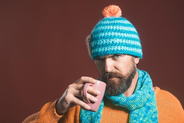 Attractive man hipster with long beard in warm hat scarf and sweater holds cup with hot beverage