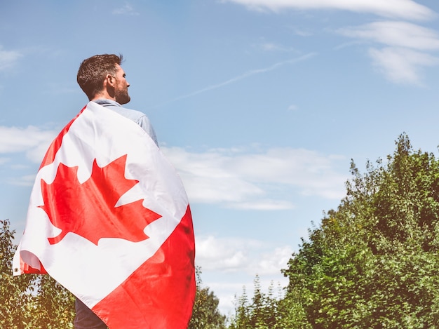 Uomo attraente che tiene la bandiera canadese sulle sue spalle sul fondo del cielo blu