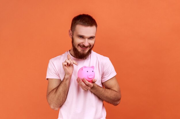 Attractive man holding pointing finger at piggy bank in hands being happy of savings