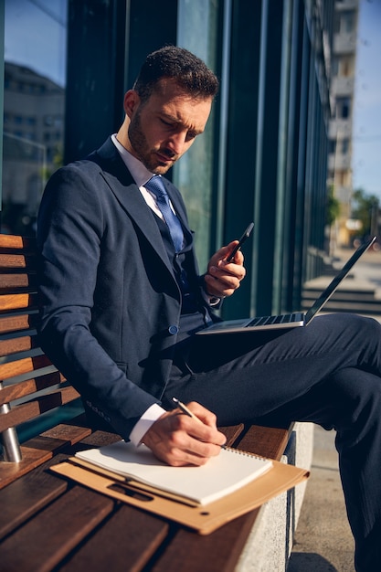 Photo attractive man sitting on bench with laptop on knees while writing and holding smartphone